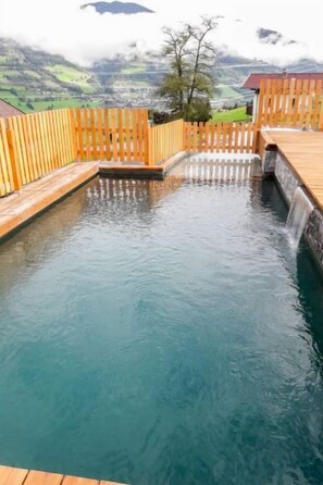 Una terrassa de fusta envoltant una piscina nítida amb una cascada, amb un paisatge de muntanyes al fons.
