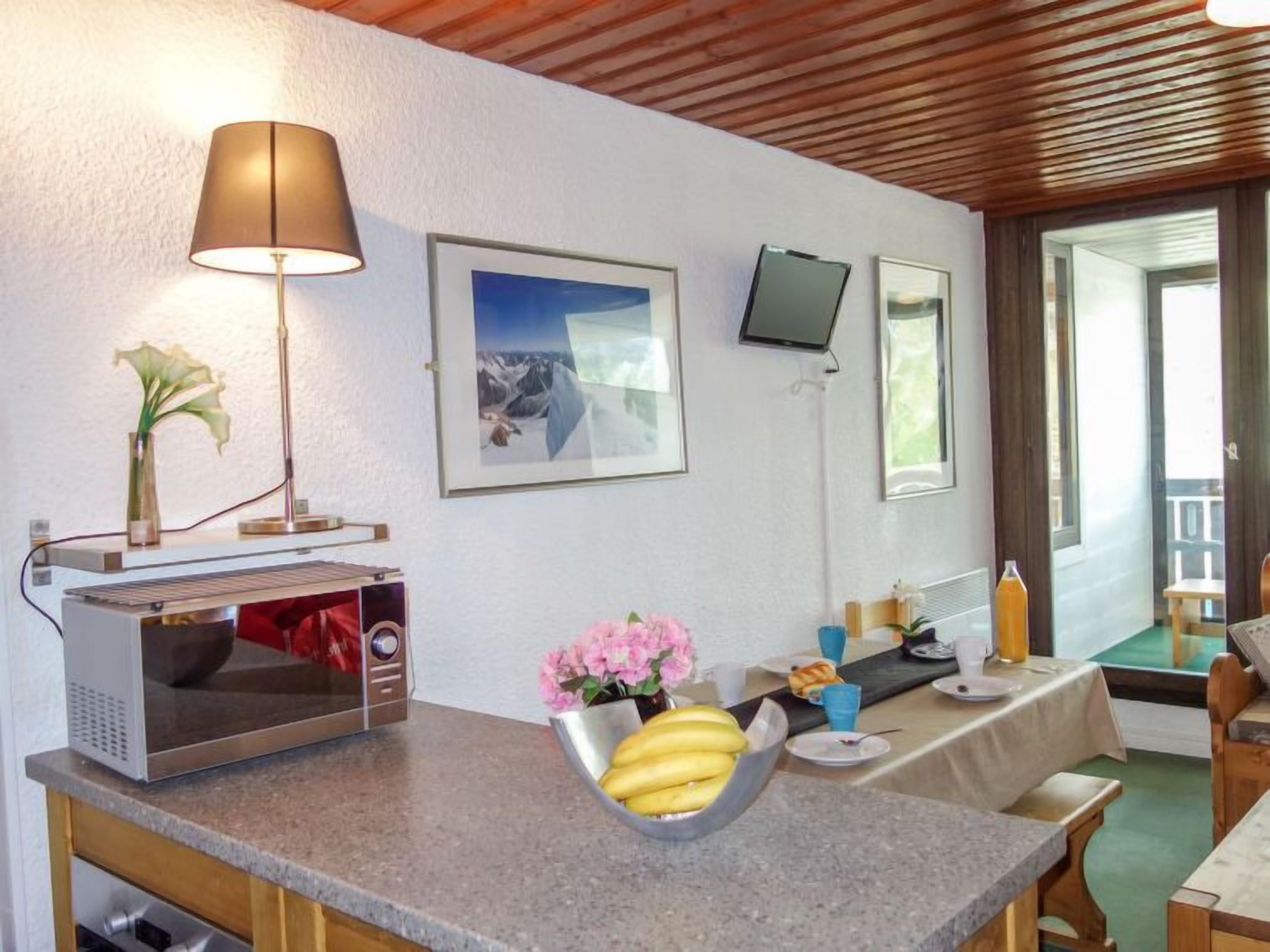 A kitchen with a microwave, a table with a bowl of fruit, a lamp, and a framed picture on the wall.