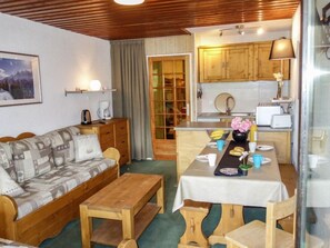 A cozy room with a wooden sofa, a wooden table with a white tablecloth, and a kitchen area with wooden cabinets and a window.