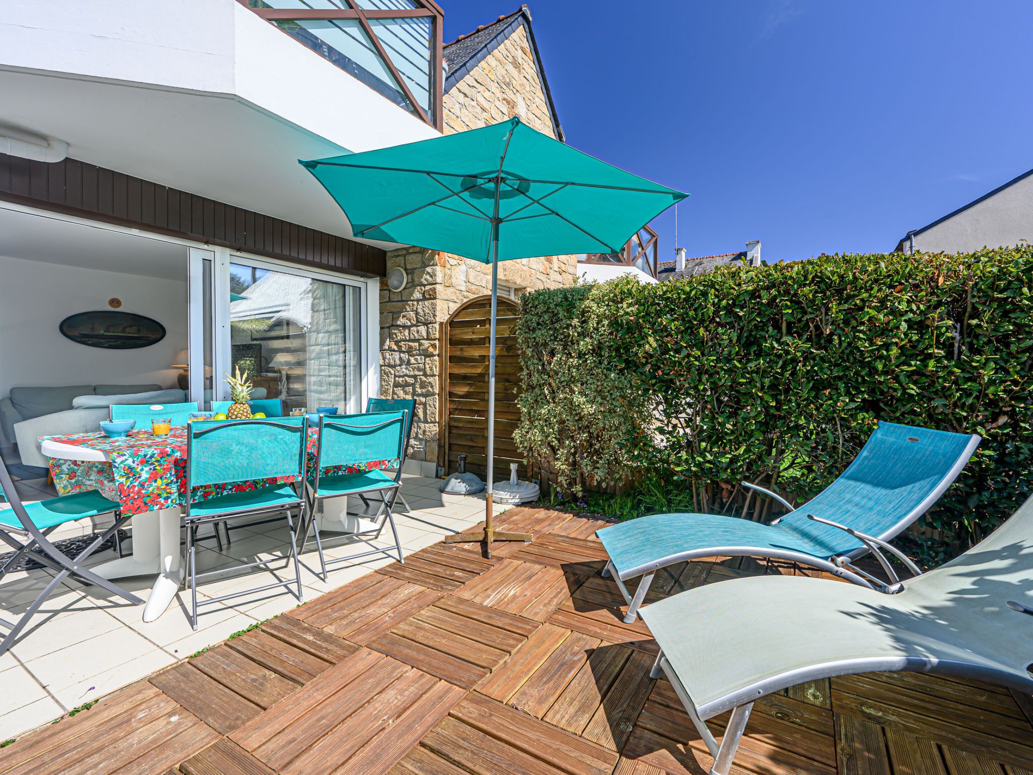 An outdoor patio with wooden flooring, a table with chairs, a large umbrella, and a hedge in the background.