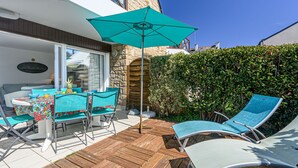 An outdoor patio with wooden flooring, a table with chairs, a large umbrella, and a hedge in the background. - Le Parc des Druides by Interhome (Carnac)