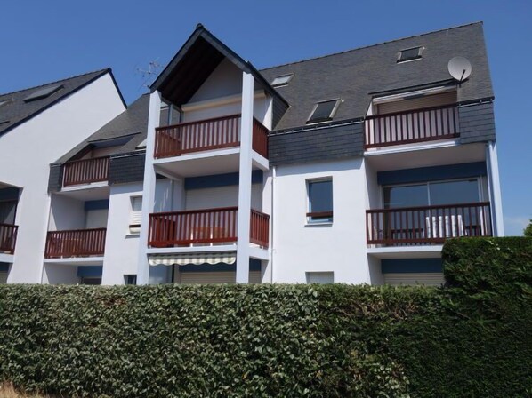 Un immeuble résidentiel à plusieurs étages, doté de balcons, d’un toit plat et d’une haie devant l’entrée.