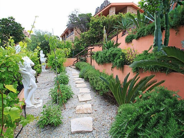 Un sentier de jardin bordé de marches en pierre, entouré d’une végétation luxuriante et de statues blanches.