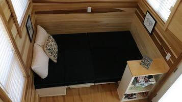Una habitación pequeña de madera con una cama, un estante con libros y una ventana con persianas.