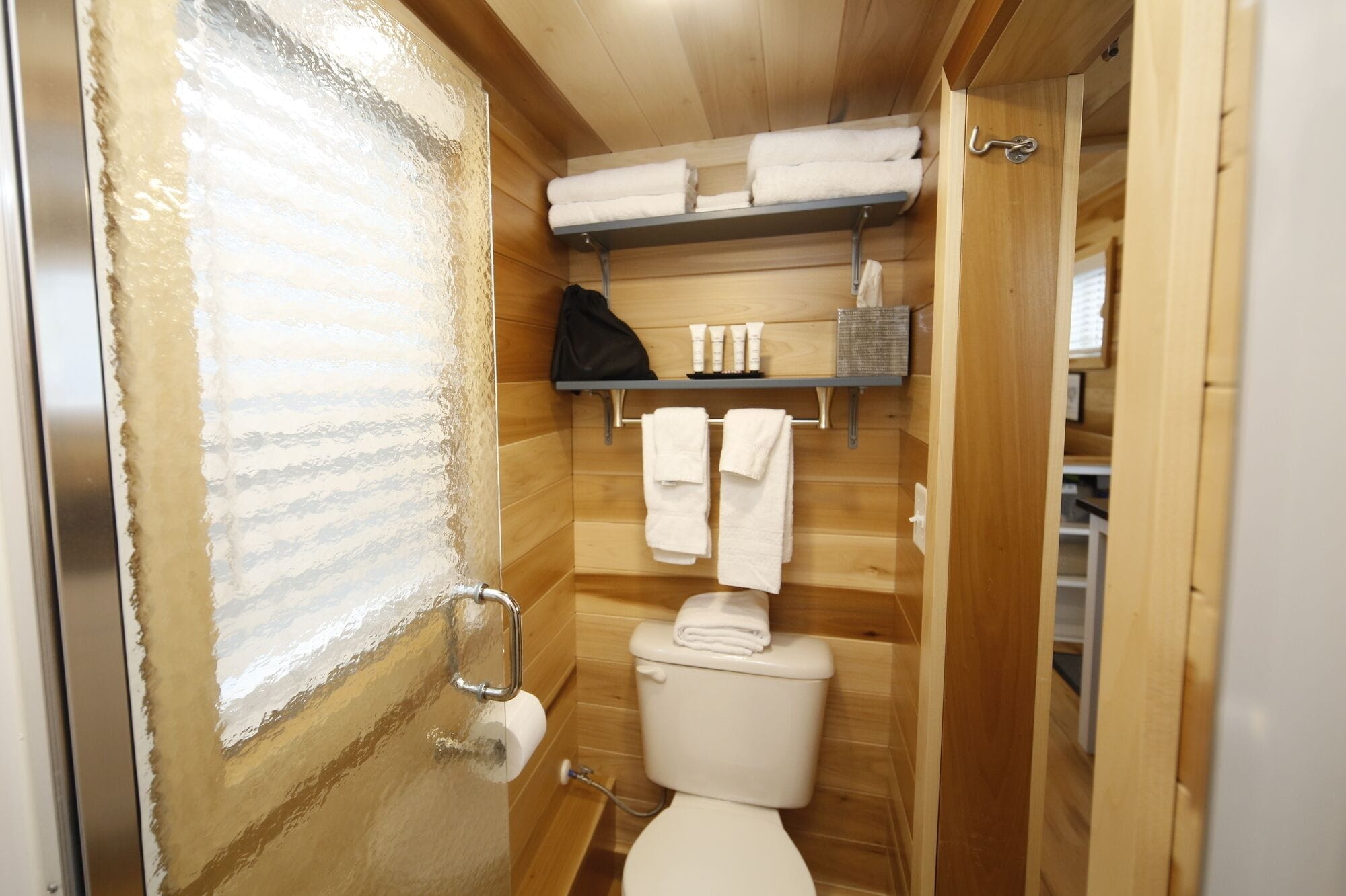A compact wooden bathroom with a toilet, a shelf with towels, and a window with frosted glass.