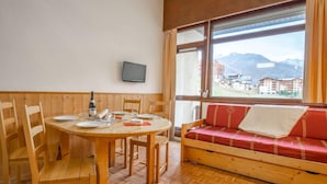 A room with a wooden dining table set for four, a red cushioned sofa, and a large window with a view of buildings and mountains. - Asters 1203 Bâtiment B4 by Interhome (Les Menuires)