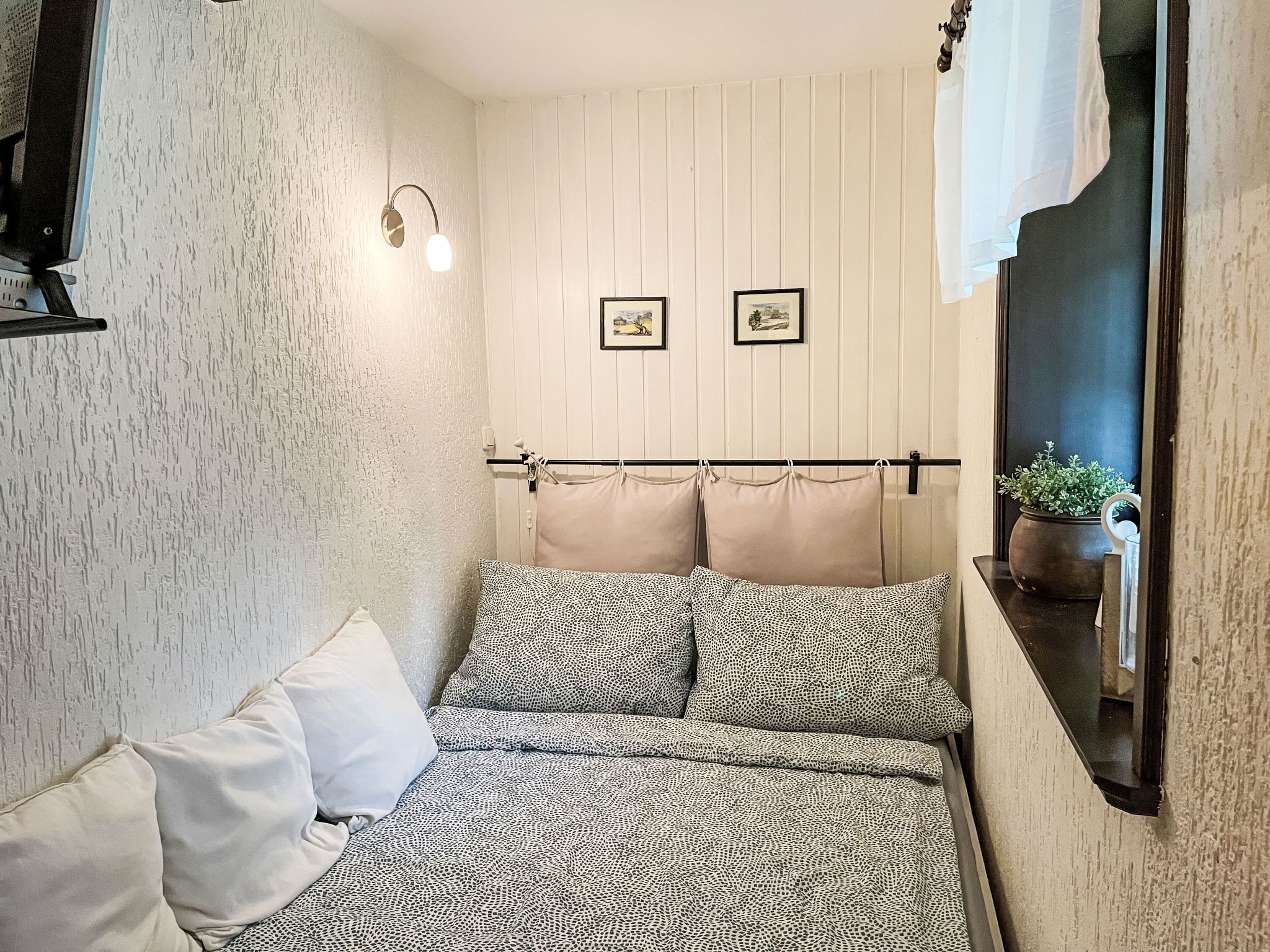 Un dormitorio con cama, almohadas, un estante con una planta y dos cuadros enmarcados.