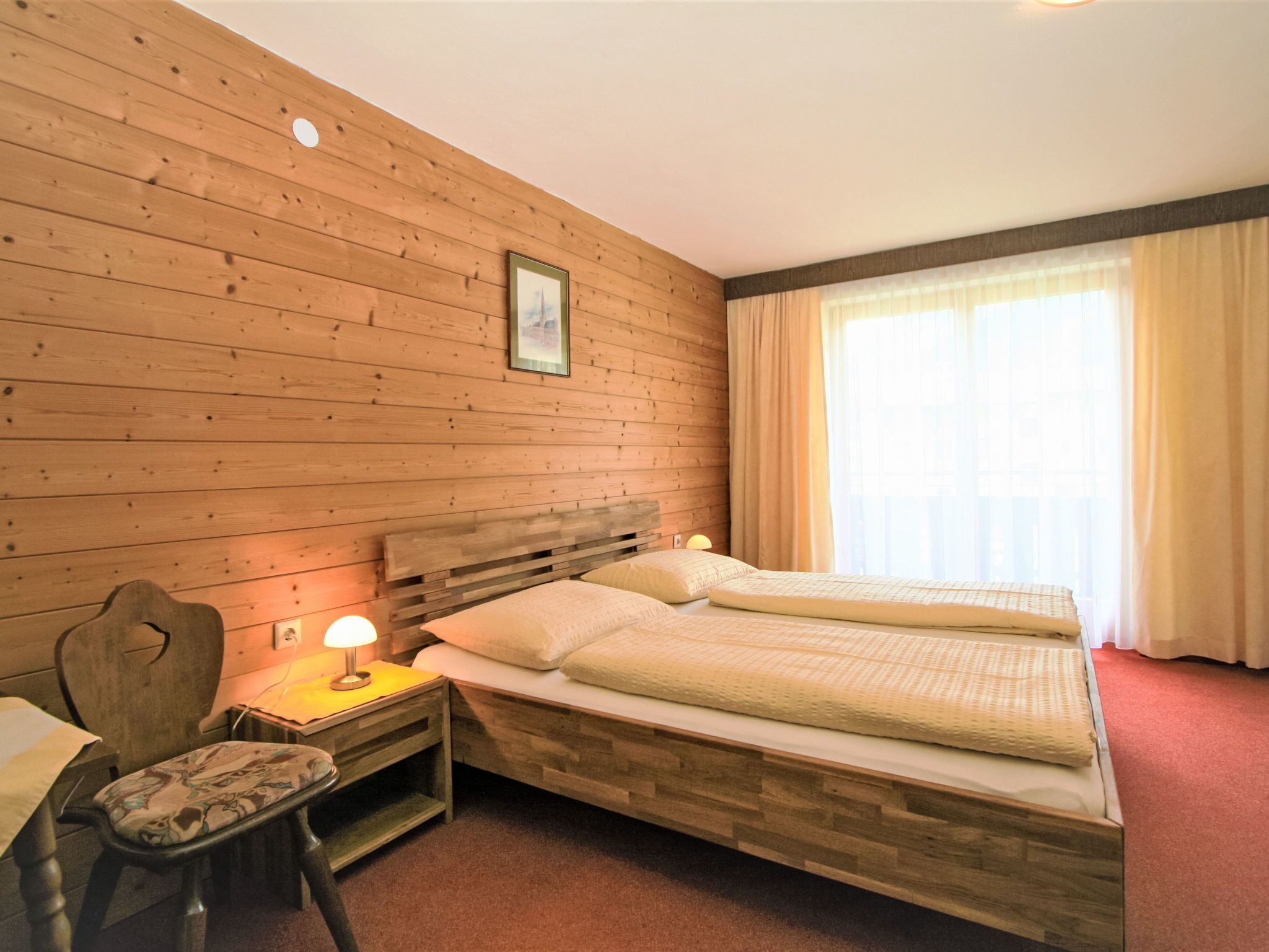 Een kamer met houten wanden, een bed met twee kussens, een nachtkastje met een lamp en een stoel.