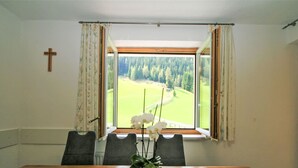 A dining room with a wooden table, two chairs, a vase with flowers, and a window with a view of a forest and golf course. - Obernberg by Interhome (Obernberg am Brenner)