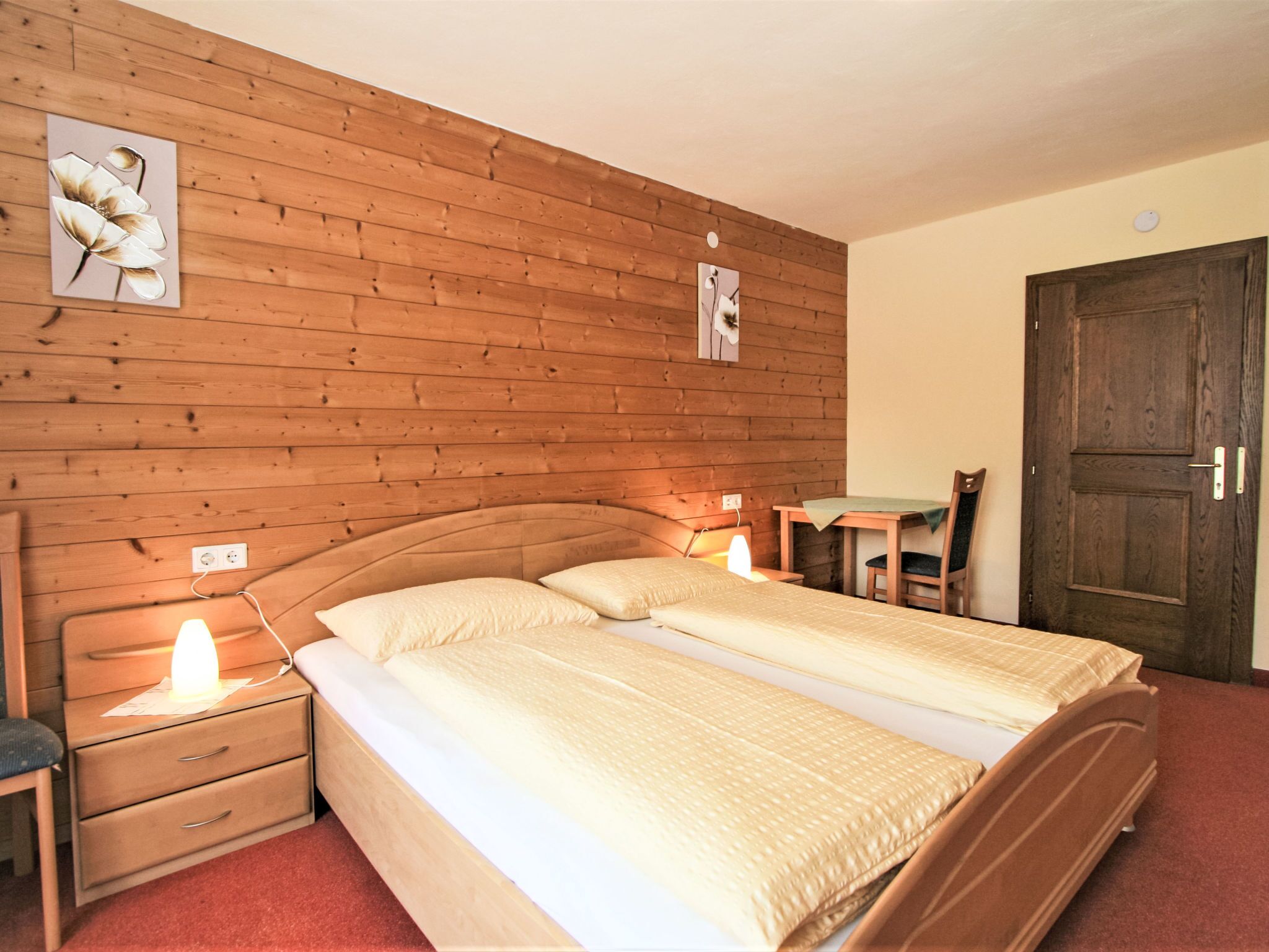 Een kamer met een houten hoofdbord, een bed met twee kussens, een nachtkastje met een lamp, een stoel en een houten deur.