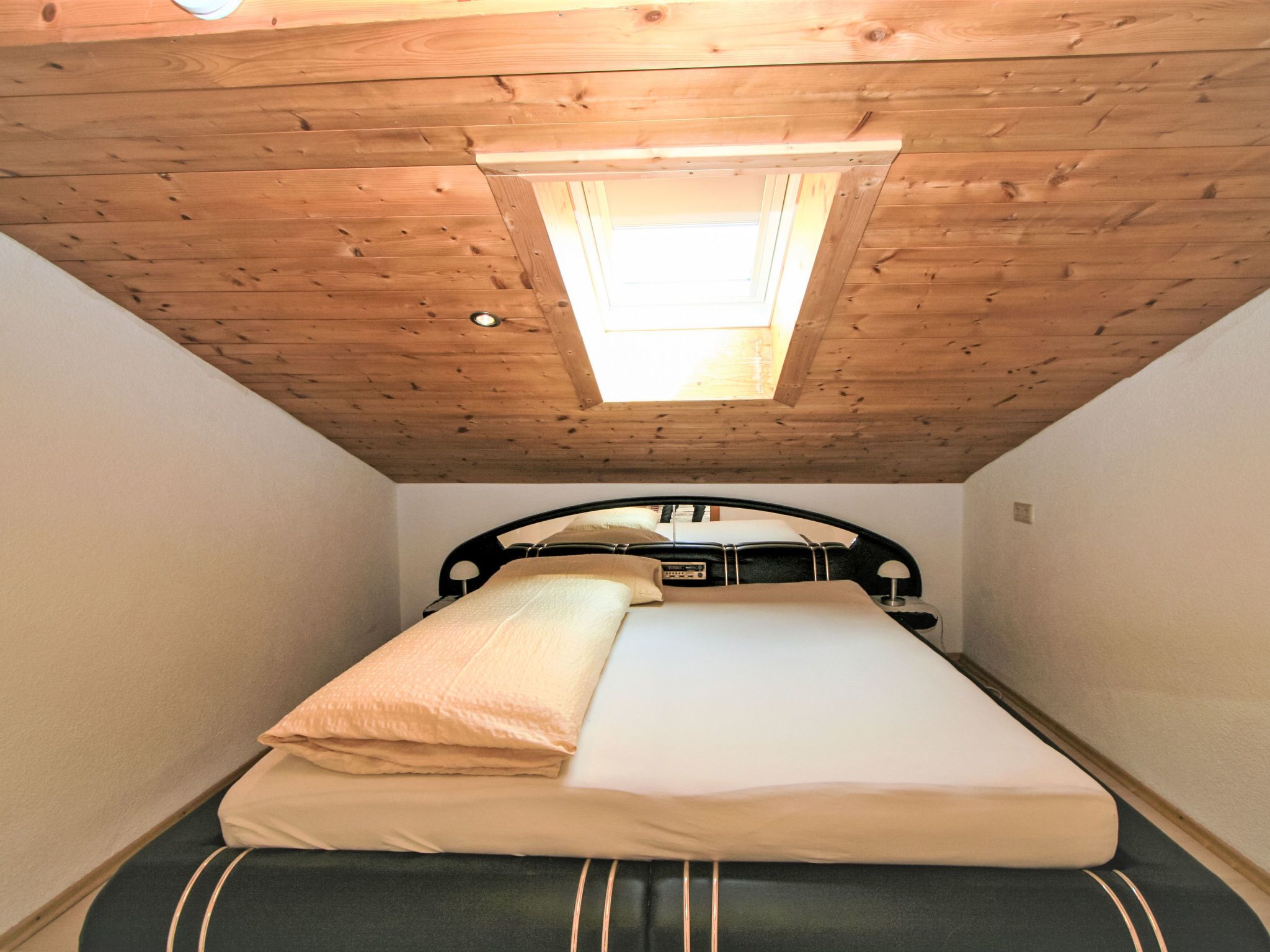 Ein kleiner, gemütlicher Raum mit schrägen Wänden und einem Oberlicht, ausgestattet mit einem Bett, einer Matratze und Kissen.