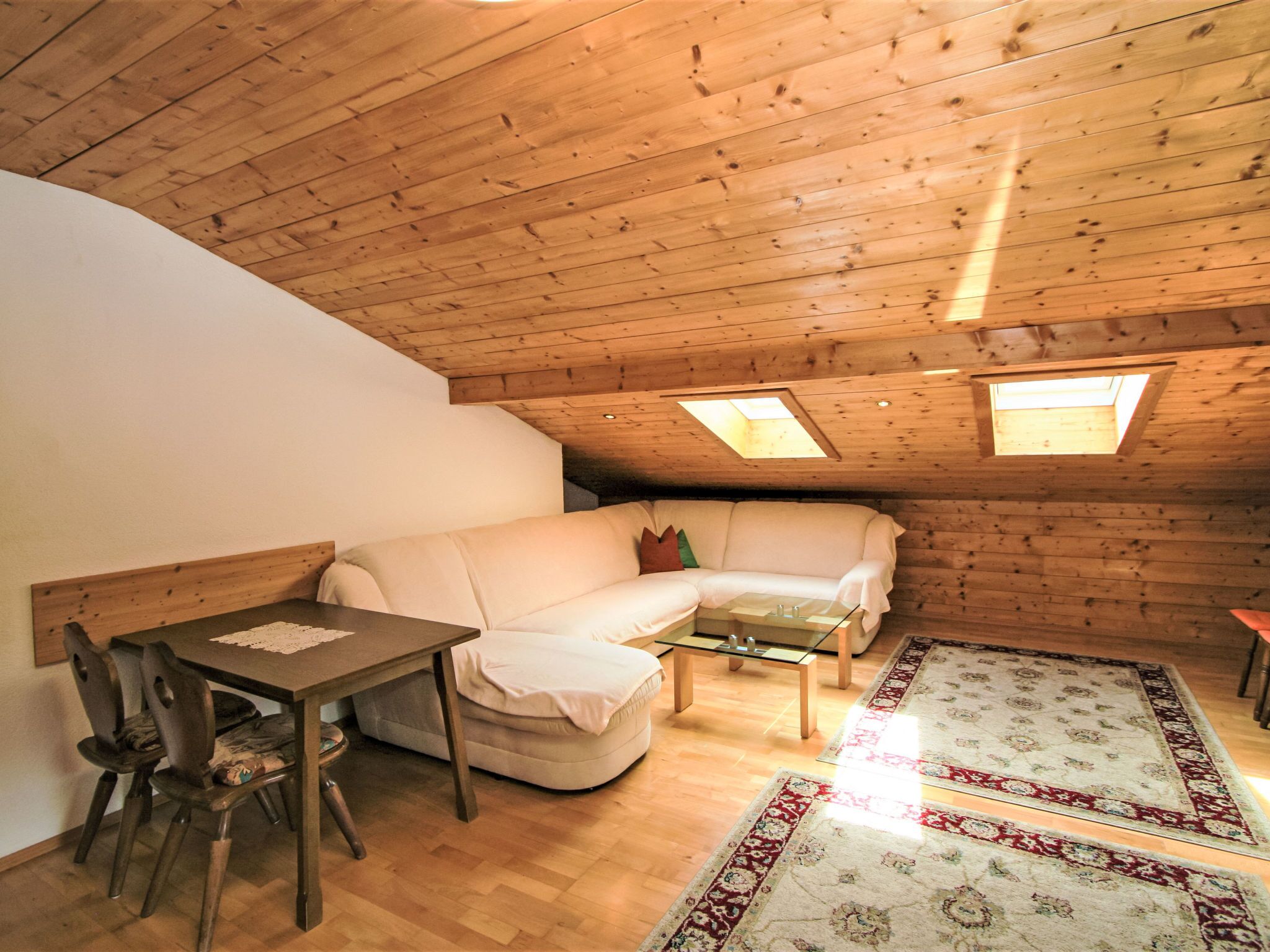 Ein gemütliches Dachzimmer mit Holzbalkendecke, einem weißen Sofa, einem Glastisch und einem gemusterten Teppich.