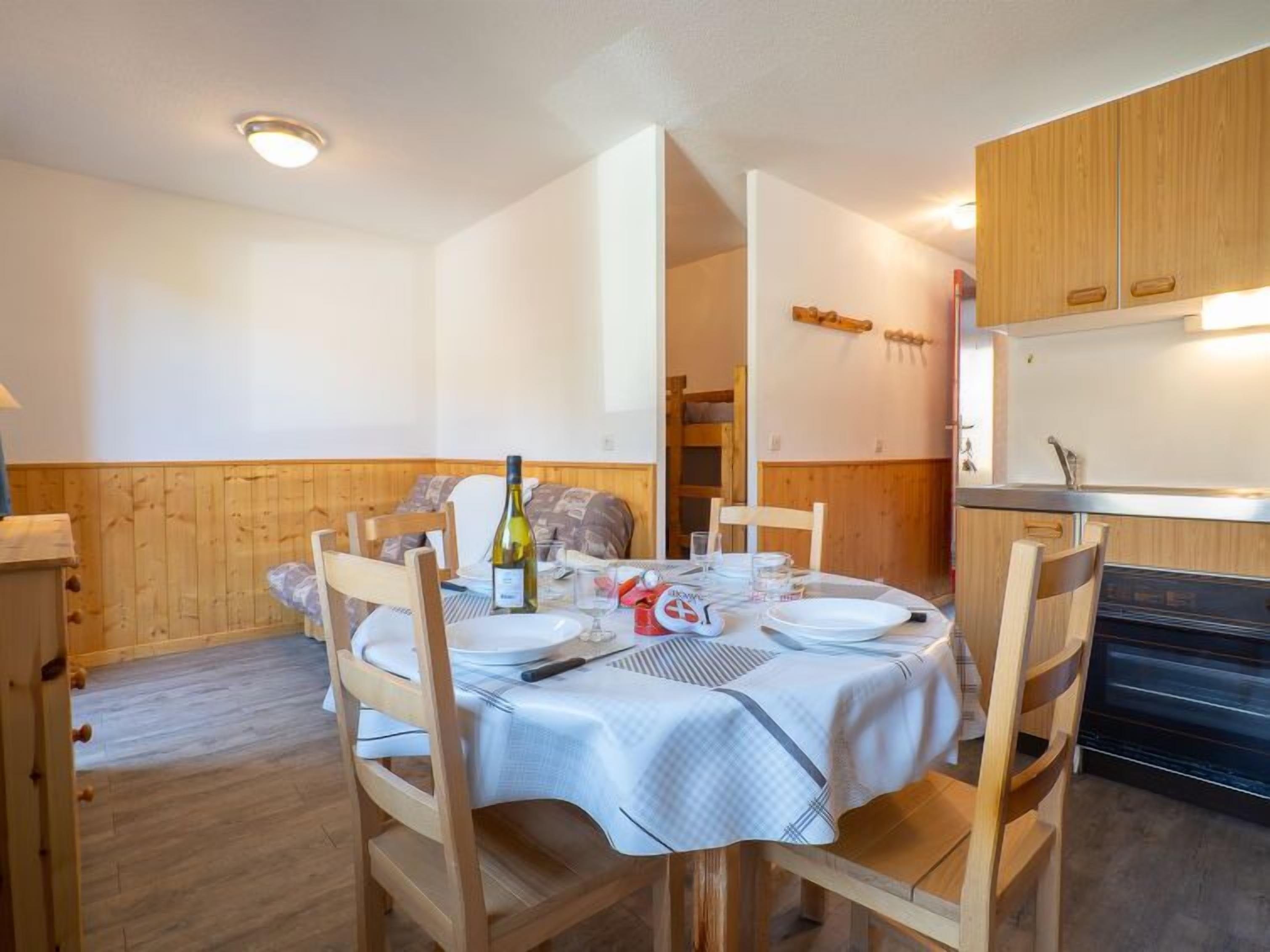 Un coin repas avec une table ronde dressée pour quatre personnes, des chaises en bois, une nappe blanche et une bouteille de vin.