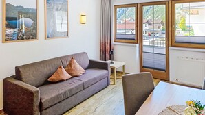 Une pièce avec un canapé gris, une petite table et une chaise. Deux tableaux encadrés ornent le mur, il y a une fenêtre avec des rideaux et, de la fenêtre, on aperçoit des bâtiments à l’extérieur.
