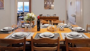 En spiseplads med et træbord dækket til et måltid, omgivet af stole. Viniglas, en vinflaske og en vase med blomster står på bordet.