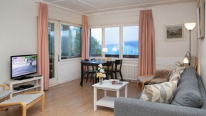 En moderne stue med en grå sofa, et fjernsyn på et stativ, et spisebord med stole og store vinduer med gardiner.