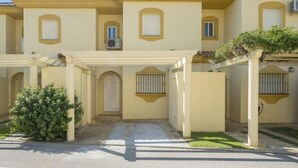 A multi-story building with a yellow facade, a covered entrance with a door, and a small garden area with a bush. - Brisamar - Novo Sancti Petri by Interhome (Chiclana de la frontera)