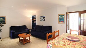 A living room with a dark blue sofa, a wooden coffee table, and a dining table with a floral tablecloth. There are bookshelves and a painting on the wall. - Brisamar - Novo Sancti Petri by Interhome (Chiclana de la frontera)