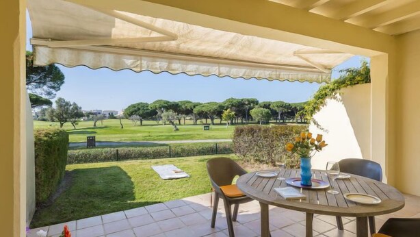A patio with a table and chairs, a view of a golf course, and a covered area with a white awning. - Brisamar - Novo Sancti Petri by Interhome (Chiclana de la frontera)