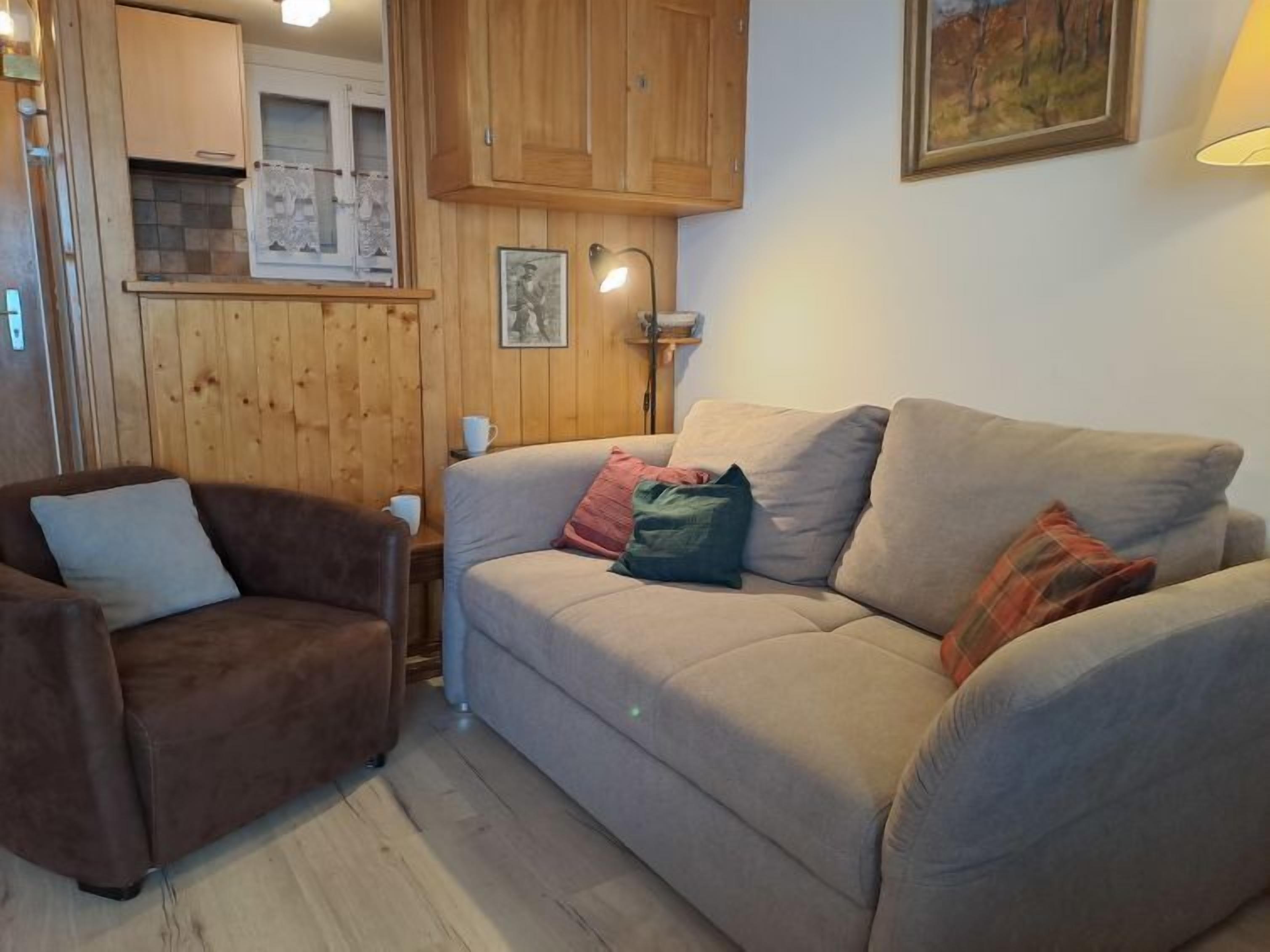 A cozy living room with a brown armchair, a beige sofa with multiple cushions, wooden paneling on the walls, and a framed picture above the sofa.