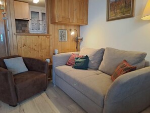 A cozy living room with a brown armchair, a beige sofa with multiple cushions, wooden paneling on the walls, and a framed picture above the sofa.