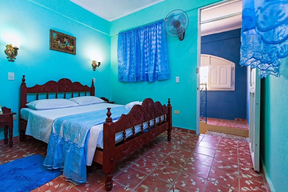 A bedroom with a wooden bed, a blue curtain, a wall-mounted fan, and a door leading to another room.