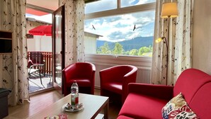 Una habitación con un sofá rojo, una mesa de madera con una botella y aperitivos, una silla roja y vistas a las montañas desde la ventana.