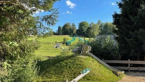 Un parque infantil con tobogán y columpios, rodeado de árboles y un césped.