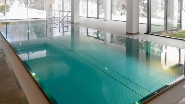 Una piscina interior con paredes de vidrio transparente, piso de baldosas y una escalera metálica que lleva a una puerta.