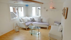 A modern living room with a white sofa, a glass coffee table, and a white armchair. There are large windows providing natural light and a view of the outside.