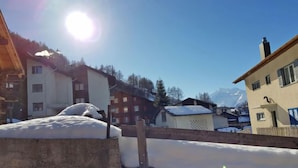 A snow-covered outdoor area with a stone wall and wooden fence, surrounded by buildings with visible windows and a mountain in the background.