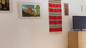 A room with a white textured wall, a framed painting of a tree, a red and black patterned cloth hanging, and a television on a wooden cabinet. - Castor und Pollux by Interhome (Täsch)
