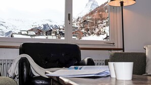Une pièce avec vue sur une montagne enneigée et des bâtiments, un fauteuil en cuir noir, une table avec une lampe et une fenêtre.