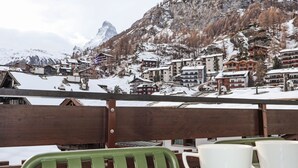 Une terrasse en bois avec des chaises vertes et des tasses blanches, d’où l’on peut admirer un village de montagne enneigé, avec ses bâtiments et un sommet imposant en arrière-plan.