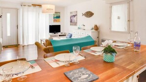 Un coin repas avec une table en bois dressée pour deux personnes, un téléviseur en arrière-plan et un lit recouvert d’une couverture turquoise.