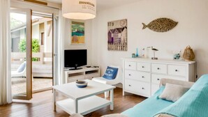 Un salon moderne avec un canapé, une table basse, un téléviseur et une commode blanche. Un décor représentant des poissons orne le mur, ainsi qu’une affiche représentant une scène de plage.