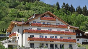 Um hotel com vários andares e varandas decoradas com flores vermelhas, com um cenário de vegetação exuberante e árvores.