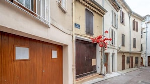 A narrow street with buildings on both sides, featuring wooden doors and shutters, and a small tree with red flowers. - Rue de la Pompe by Interhome (Antibes)