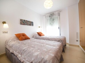 A bedroom with two beds, a wall-mounted light, a window with sheer curtains, and a door with a vent.