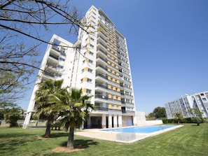 A high-rise residential building with multiple balconies, a swimming pool, and a well-maintained lawn with palm trees.