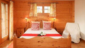 A wooden-paneled bedroom with a bed, two bedside tables, a window with curtains, and a chair covered with a white cloth. - Boule de Neige by Interhome (Verbier)