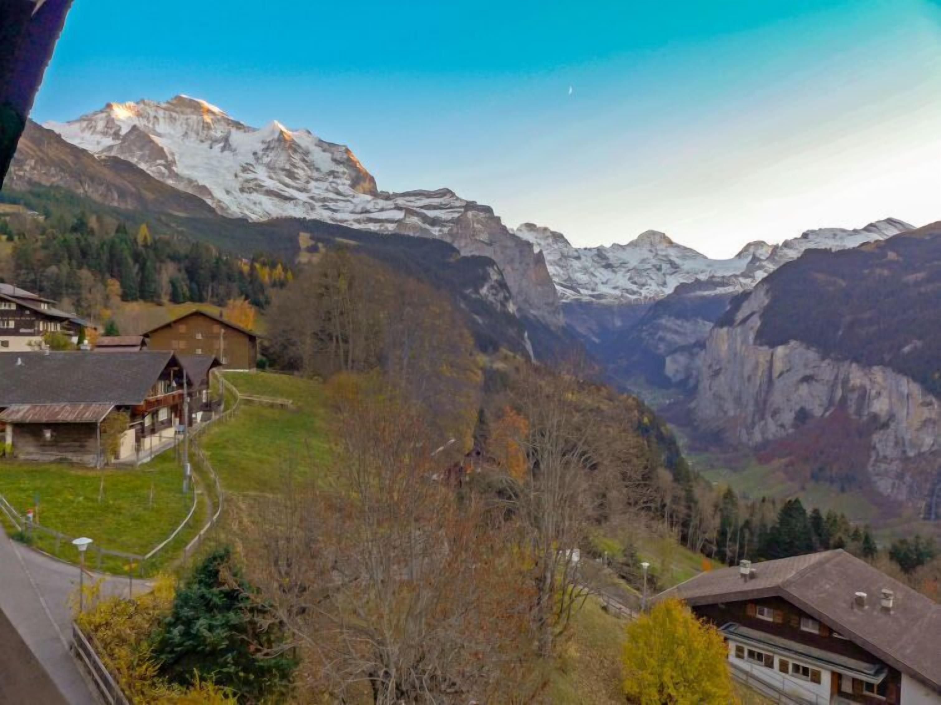 Ein Bergdorf mit traditionellen Holzhäusern, freier Aussicht auf schneebedeckte Gipfel und einem Tal darunter.