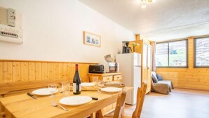 Une table de salle à manger en bois, mise avec des assiettes, des couverts et une bouteille de vin. On y trouve également un four à micro-ondes, un réfrigérateur et une fenêtre donnant sur des immeubles.