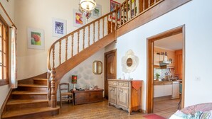 Un escalier en bois menant à un deuxième étage, un coin repas avec une table et des chaises, et une cuisine visible à travers une porte ouverte.