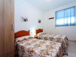 Een kamer met twee bedden, een houten garderobe, een raam met blauwe gordijnen en een wandplank met plantenpotten.