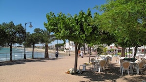 Um calçadão pavimentado com mesas ao ar livre, uma praia ao fundo e céu claro.