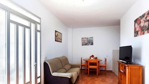 Uma sala de estar com sofá, mesa de jantar e cadeiras, TV sobre um suporte de madeira e uma janela com persianas.