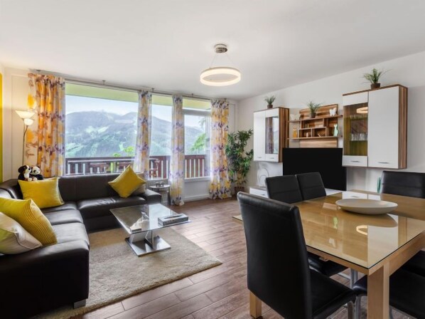 A modern living room with a sofa, coffee table, and dining table. There is a balcony with a view of the mountains.