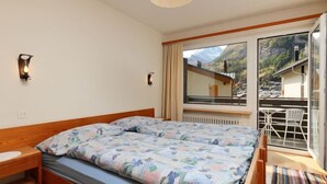 A bedroom with a bed, bedside tables, a window with a mountain view, and a balcony with chairs. - Apartment Apollo by Interhome (Zermatt)