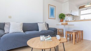 A modern living room with a grey sofa, a round wooden table, and a kitchen area with a window. - Residencial Gran Sol by Interhome (L'Escala)